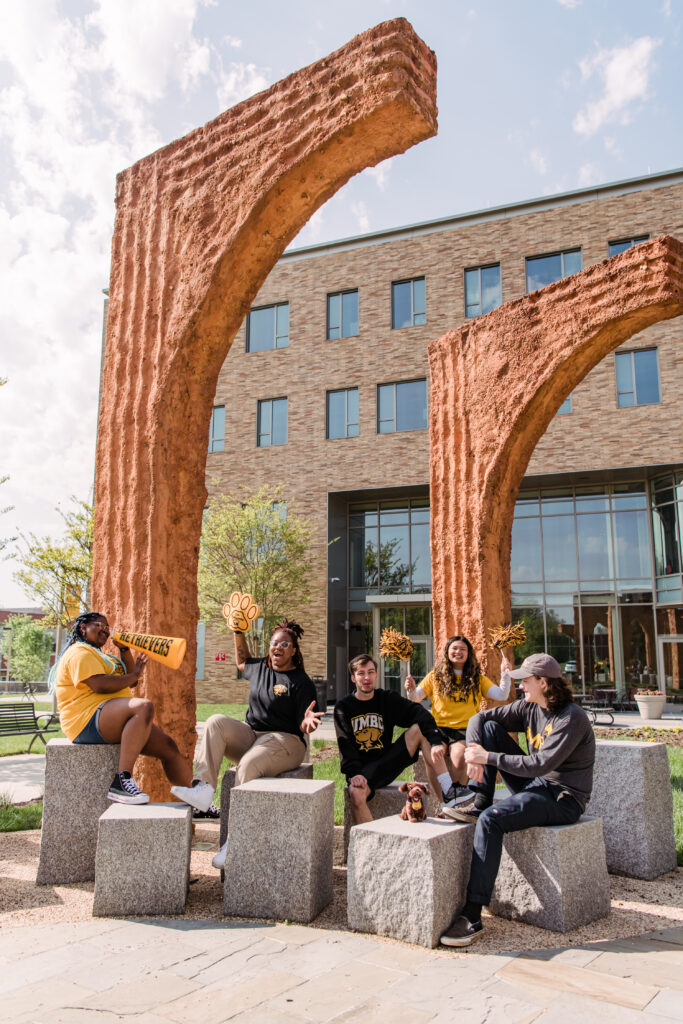 Excited students dressed in UMBC apparel sitting under the sculptures in front of the PAHB building