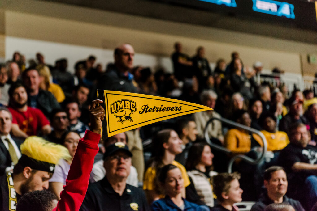 Students cheering at a basketball game. Photo centered on a student holding up a UMBC Retrievers pennant.