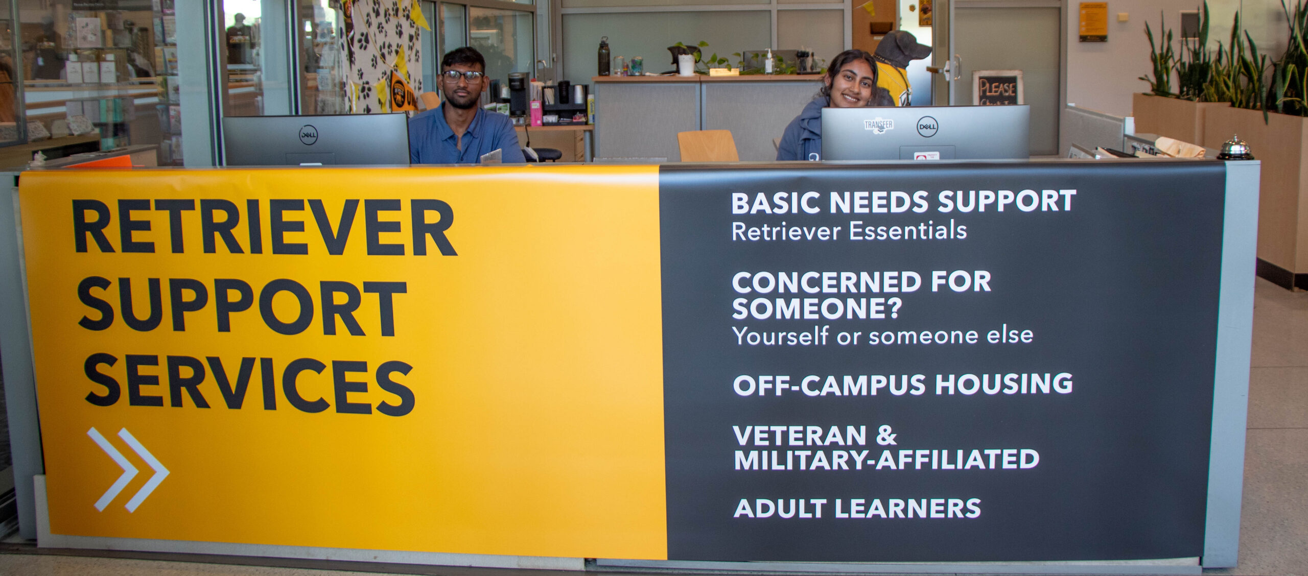 Two friendly students sitting behind the Retriever Support Services desk down in the Main Street Lounge.