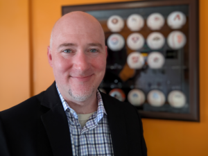 Simon Reilly standing in front of an orange wall with a baseball display case in the background.